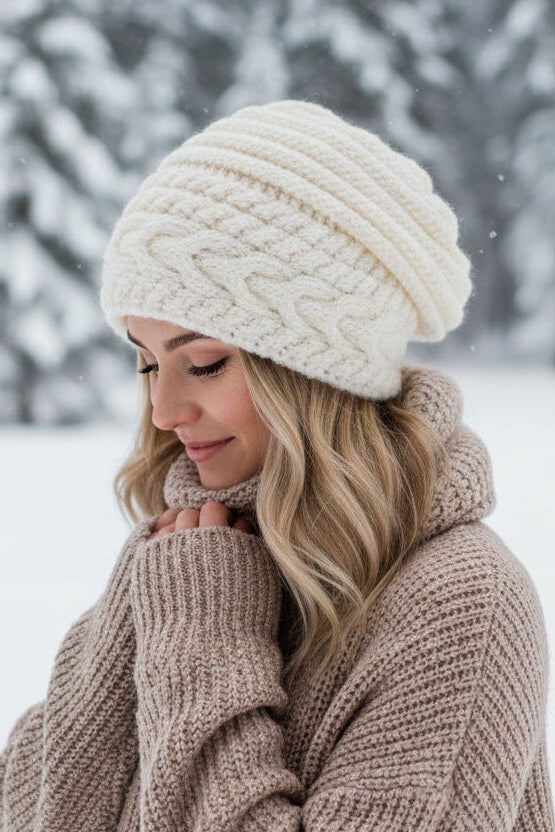 Cream cable-knit slouchy beanie hat displayed on a woman's head in a winter scenery.