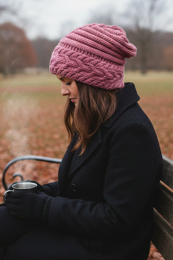 A dusty rose chunky knit slouchy beanie hat with cable and ribbed patterns displayed on a woman's head sitting on the bench in the park.