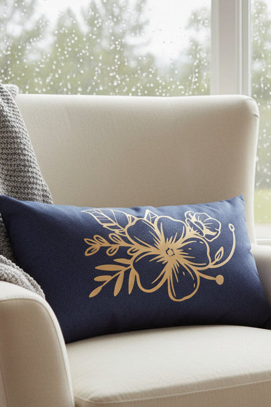 A rectangular navy blue pillow with a gold floral vinyl design on the front, displayed on an armchair by the window.